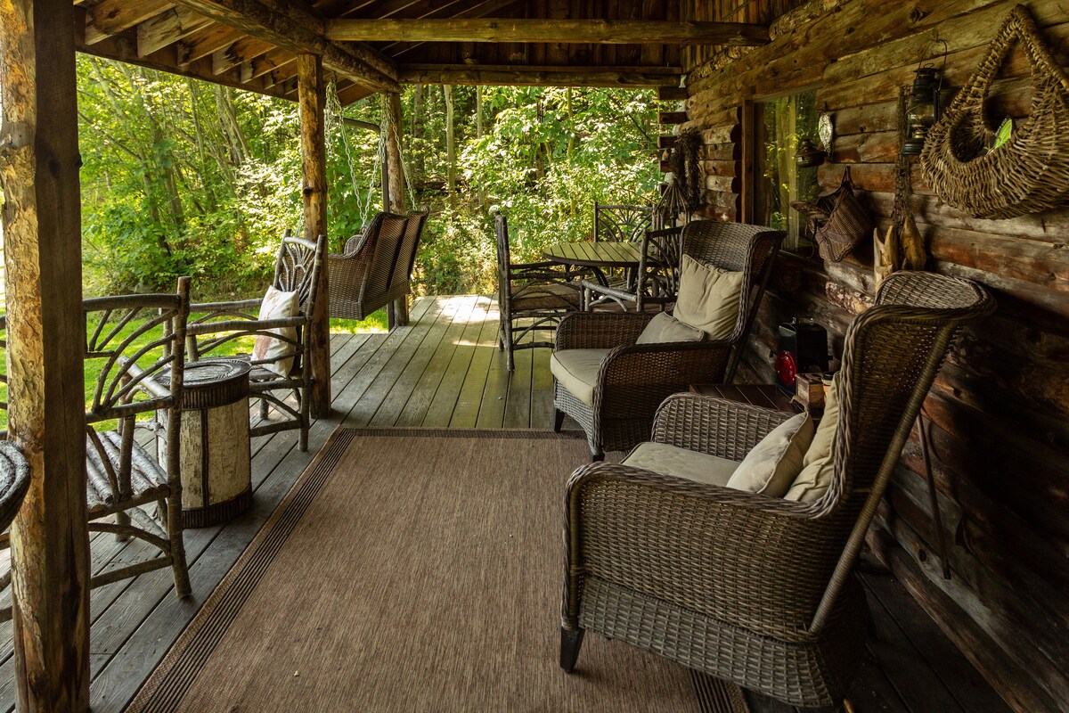 A comfortable front porch is displayed, featuring several wicker chairs and a small table. The space is surrounded by green foliage, providing a serene view of the natural environment. A swing chair adds a relaxing element, while the wooden deck enhances the rustic charm.