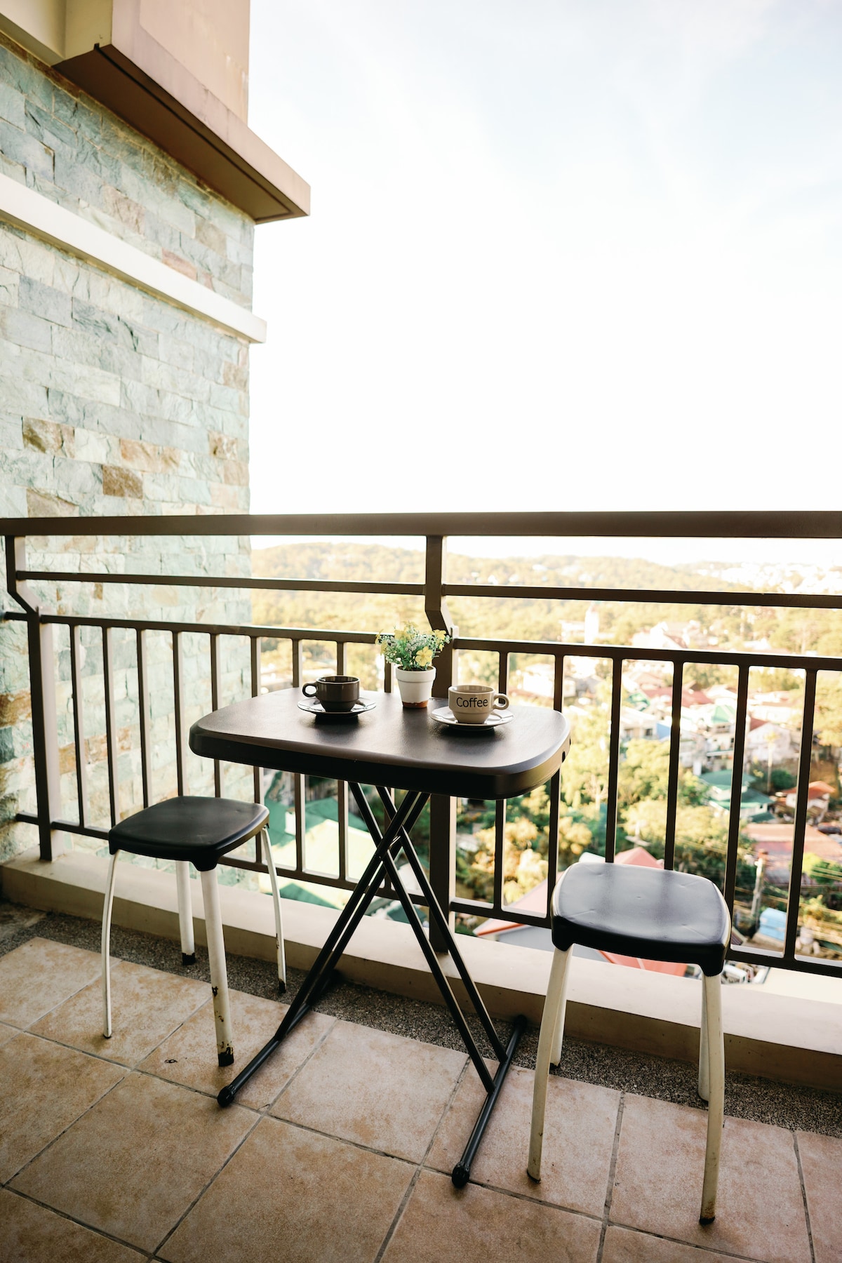 A small balcony is furnished with a simple black table and two chairs. On the table, two mugs are placed beside a potted plant. The stone wall provides a contrasting backdrop, while a scenic view of the mountains and city can be seen beyond the railing.