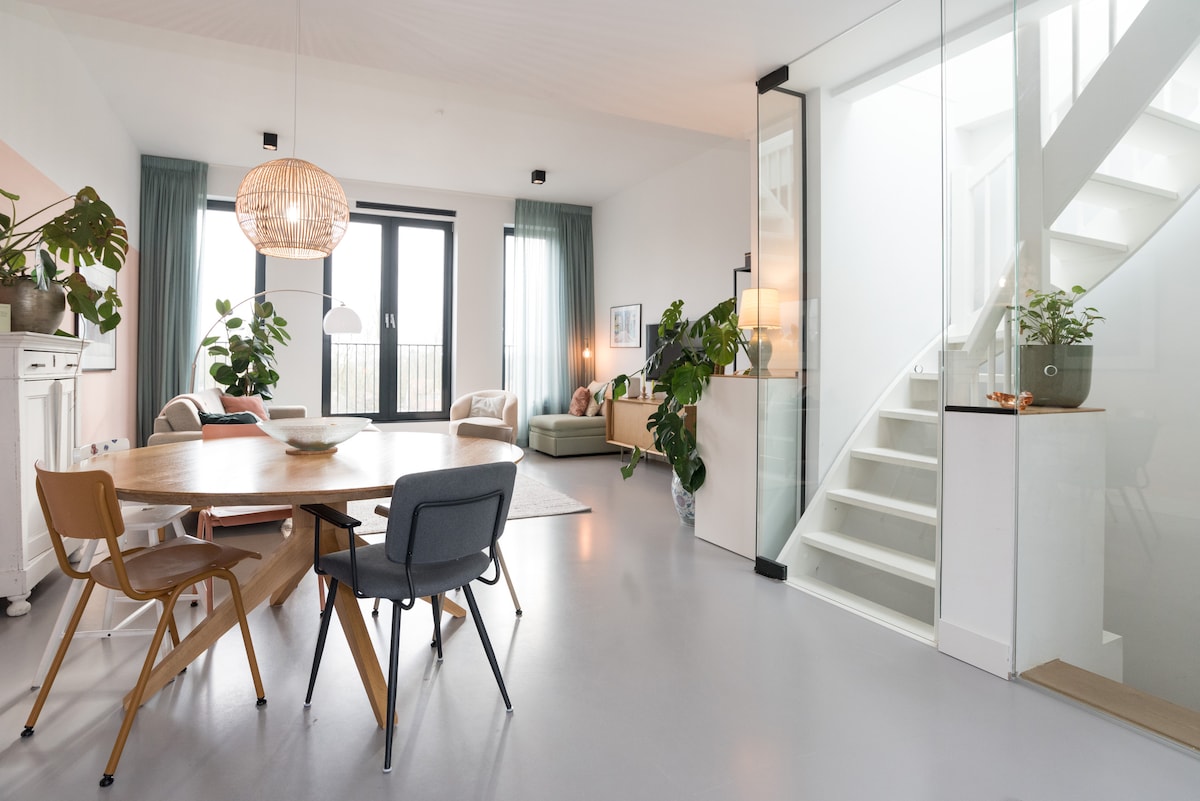 A bright, open living space features a dining area with a round wooden table surrounded by chairs. Large windows provide natural light, while potted plants add greenery. A cozy seating area is visible, complemented by soft furnishings and an elegant staircase leads to the upper level.