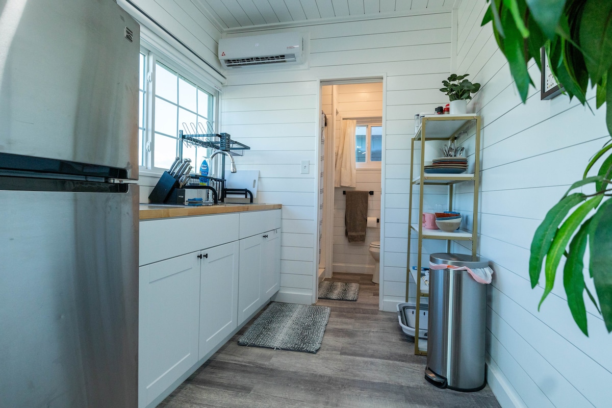 A modern kitchen area is presented with white cabinetry and a wooden countertop. Essential appliances, including a refrigerator and a microwave, are visible. A pantry shelf holds various kitchen items, while a small rug adds comfort underfoot. Natural light pours in through the window.