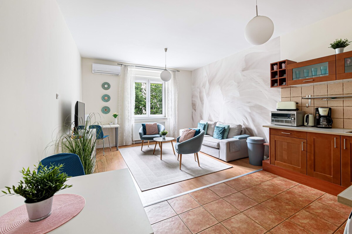 A light-filled living space features a comfortable sofa with teal cushions, accompanied by two blue chairs and a dining table. A large window provides views of greenery, while a wall mural adds an elegant touch. The kitchen area is equipped with wooden cabinets and modern appliances.