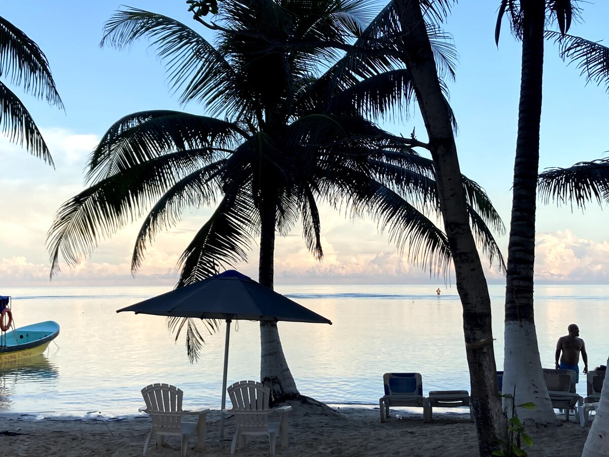 Gentle waves lap at a sandy shore under a soft sunrise. A shaded umbrella stands over white lounge chairs, surrounded by palm trees swaying in the breeze. Silhouetted figures enjoy the serene beachfront, with a boat anchored in the calm water.