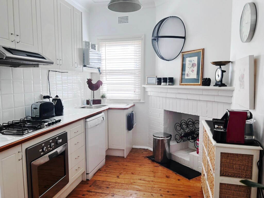 A well-appointed kitchen features white cabinetry and a wooden countertop. Modern appliances include a stovetop, oven, and dishwasher. Natural light filters through a set of horizontal blinds, illuminating a decorative wall clock and artwork positioned on the fireplace mantel.