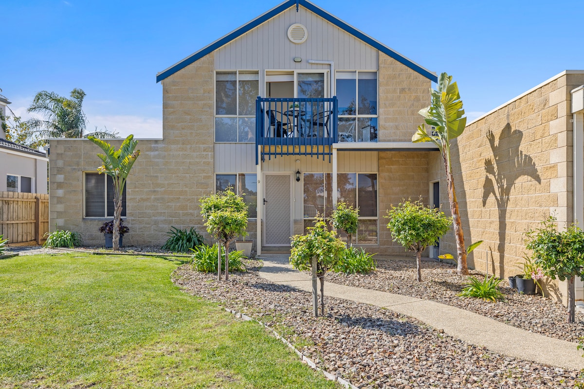 The exterior of a two-story stone building is displayed, featuring a private balcony on the upper level. Well-maintained landscaping includes green grass, shrubs, and palm trees, creating a welcoming entryway with a pathway leading to the front door.
