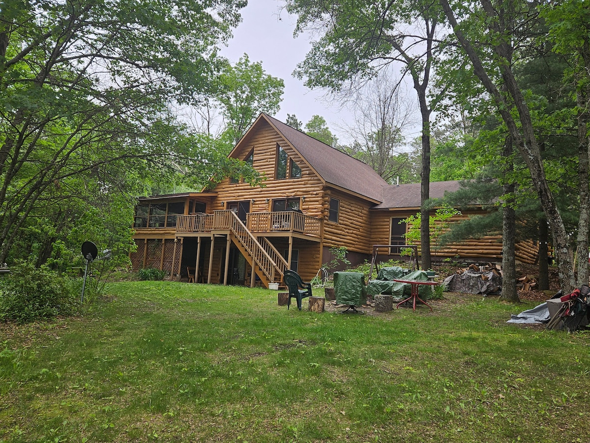 The exterior of a rustic log cabin features a wide staircase leading to an upper deck, surrounded by lush green trees. A spacious yard is visible, along with outdoor seating areas and covered equipment. The inviting log walls reflect a connection to nature.