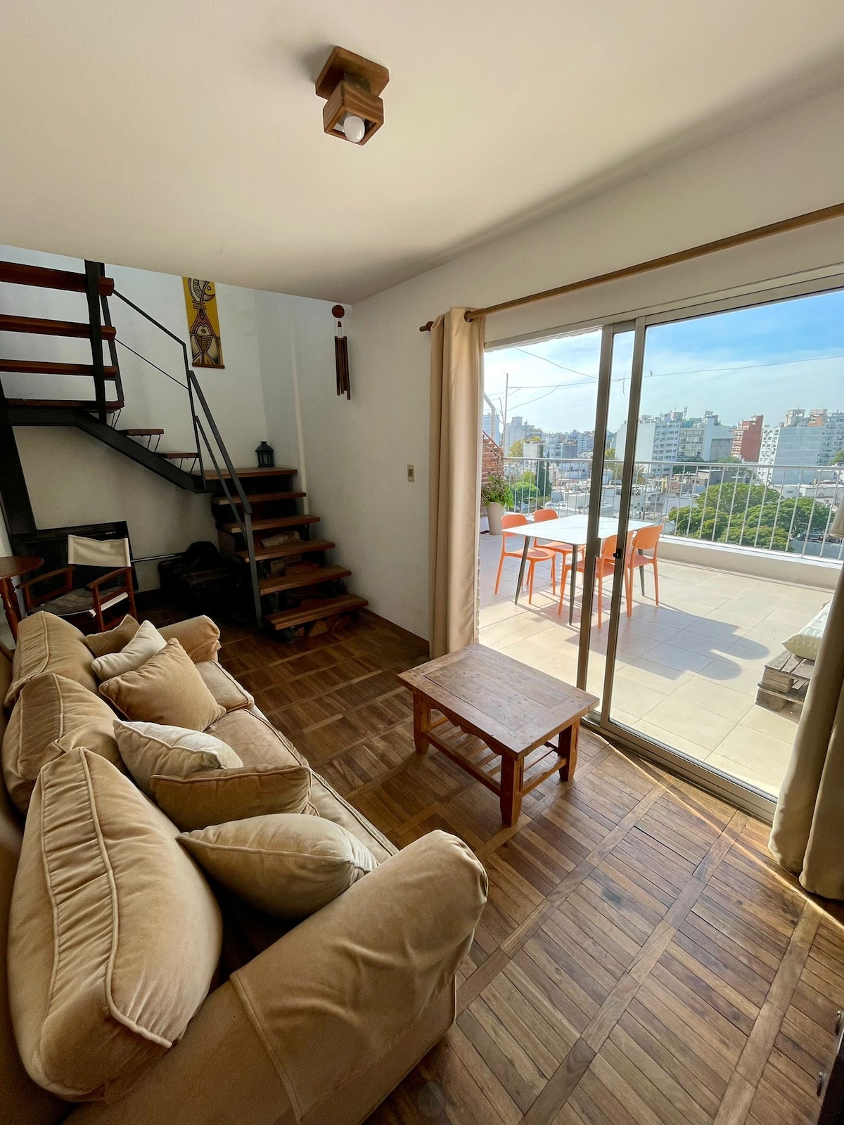 The living area features a comfortable sofa set arranged around a wooden coffee table. A large glass sliding door provides access to the terrace, offering views of the city. Natural light fills the space, accentuating the wooden flooring and the staircase leading to the upper level.