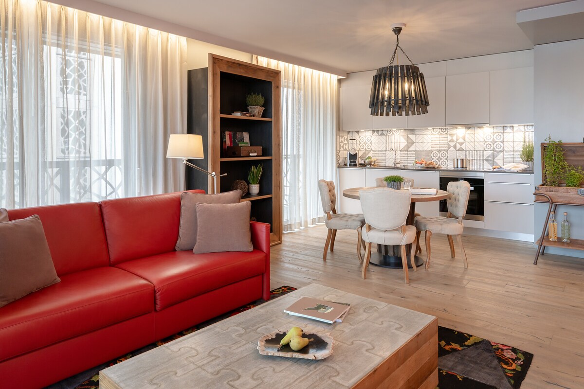 A living area is furnished with a red sofa and decorative pillows, complemented by a wood bookcase. A round dining table with upholstered chairs is positioned nearby. Natural light filters through sheer curtains, illuminating the space and highlighting the stylish kitchen area in the background.
