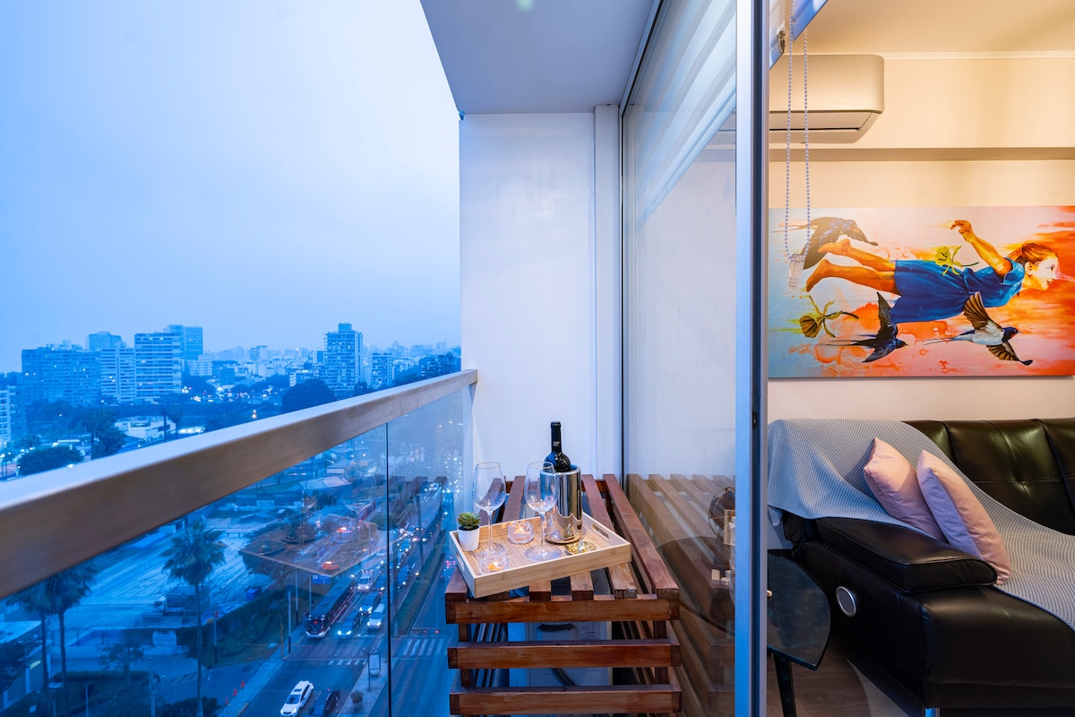 A private balcony features a wooden table set with a bottle of wine and glasses, offering panoramic views of the city. A vibrant piece of artwork adorns the wall, contributing to a cozy atmosphere. Soft lighting creates an inviting space to relax.