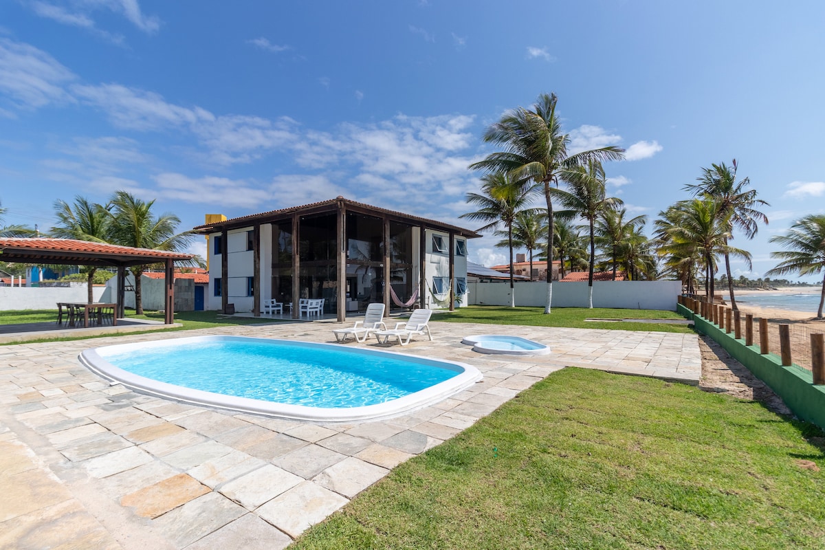 A spacious outdoor area is showcased, featuring a large pool surrounded by lounge chairs. Palm trees dot the landscape, and a two-story house is visible in the background. The area offers an unobstructed view of the coastline and a sunny blue sky.