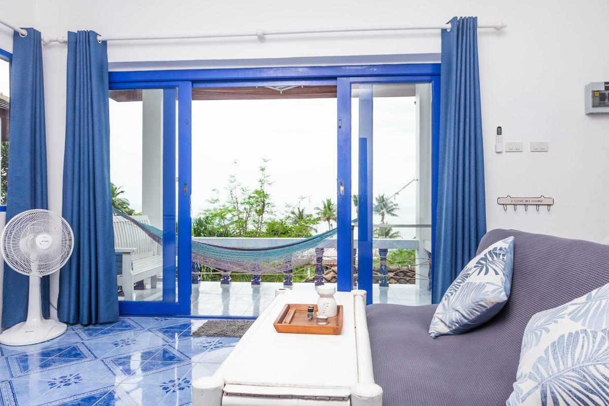 A bright living space features a cozy sofa adorned with decorative cushions and a wooden tray on a coffee table. Large sliding glass doors lead to a balcony with a hammock, offering views of lush greenery and the outdoor area.