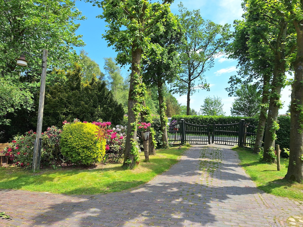 A gravel driveway leads to a wooden gate, framed by lush greenery and blooming flower bushes. Sunlight filters through the trees, highlighting the vibrant colors of the garden. The peaceful surrounding nature enhances the serene entrance to the property.