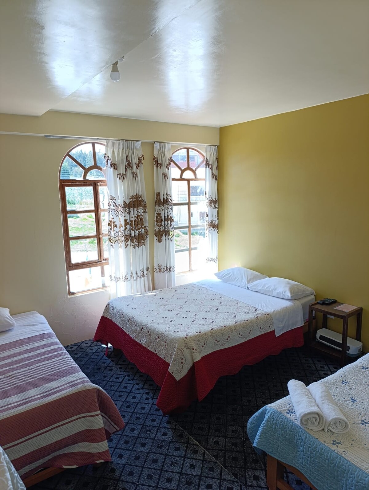 A well-lit room features two beds, one with a decorative red bedspread and the other with a patterned cover. Large windows with sheer curtains allow natural light to fill the space. A nightstand stands beside the bed, complemented by neatly arranged towels.