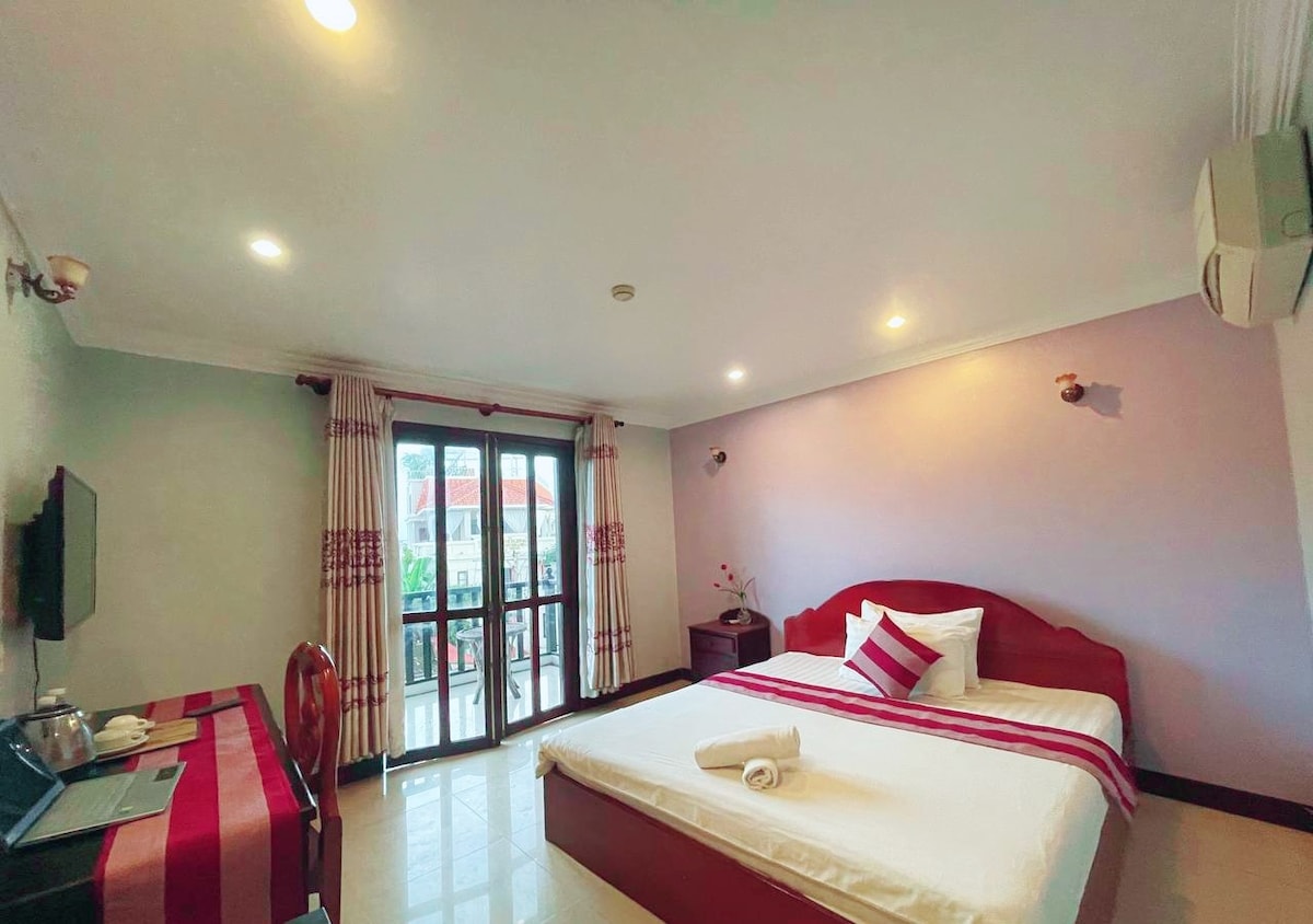 A spacious bedroom features a large bed with a neatly rolled towel placed on top. A flat-screen TV is mounted on the wall opposite the bed, and a small table with chairs is seen in the corner. Natural light enters through sliding doors leading to a balcony.