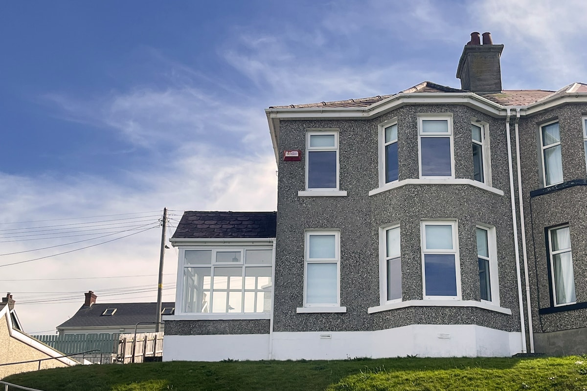 A modern three-story house with a grey stone exterior is shown against a bright blue sky. Large bay windows on the front reflect natural light. A grassy area borders the house, creating a serene outdoor space.