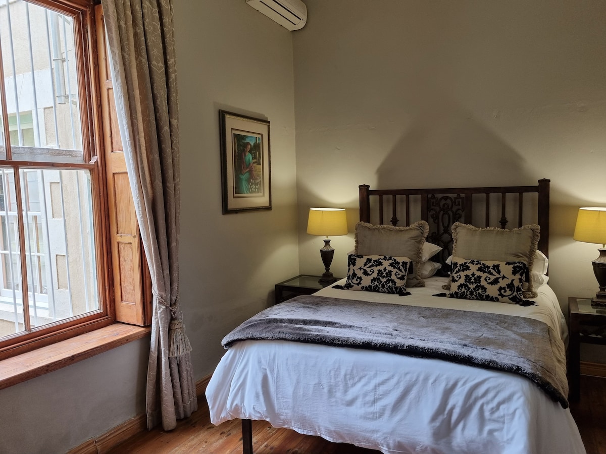 A comfortable double room features a wooden bed framed with decorative pillows. Two bedside lamps provide soft lighting. A large window reveals natural light, framed by elegant curtains, while a piece of art decorates the wall, complementing the wooden flooring.