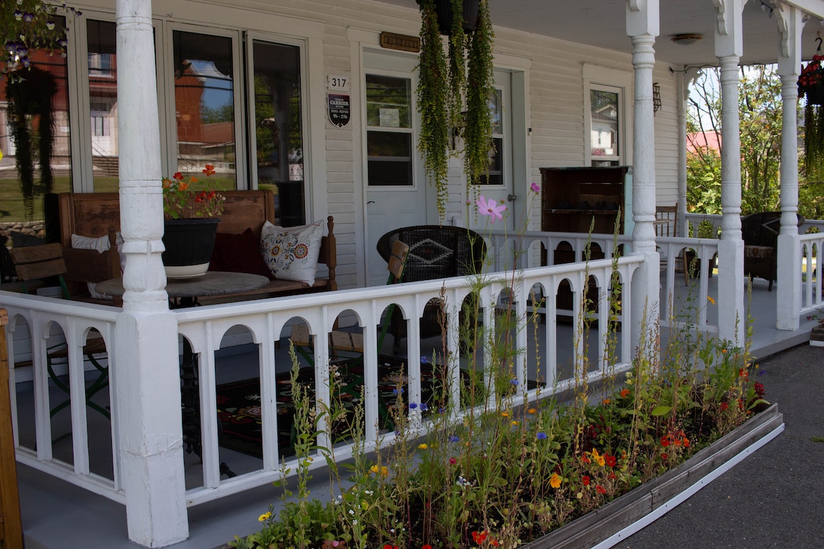 A charming porch features several wooden chairs and tables arranged for relaxation. Lush greenery and colorful flowers are integrated into the flower beds, enhancing the welcoming ambiance. The front door, framed by windows, invites guests into the heritage home with vintage character.