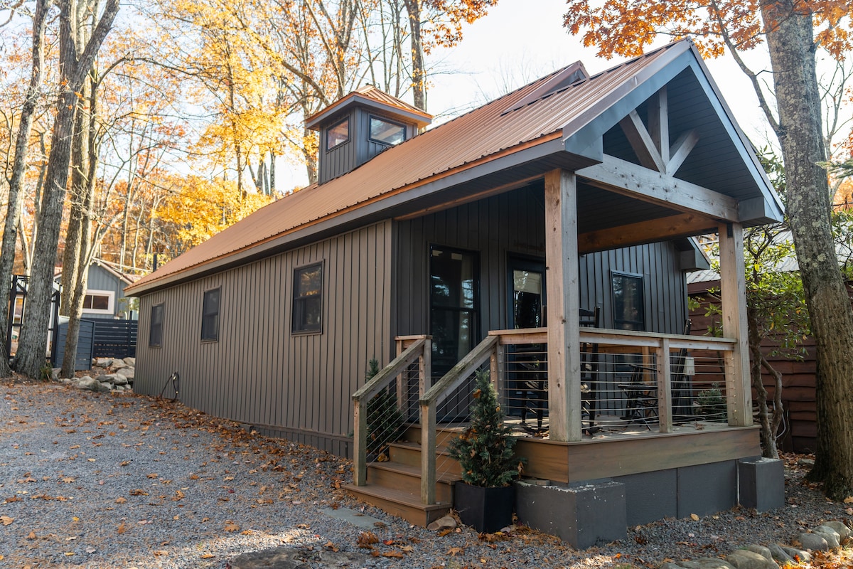 The exterior of the tiny house is presented, featuring a distinctive copper roof and a cupola. The entrance is framed by a welcoming porch with wooden railings. Surrounding trees display autumn foliage, complementing the home’s rustic charm.