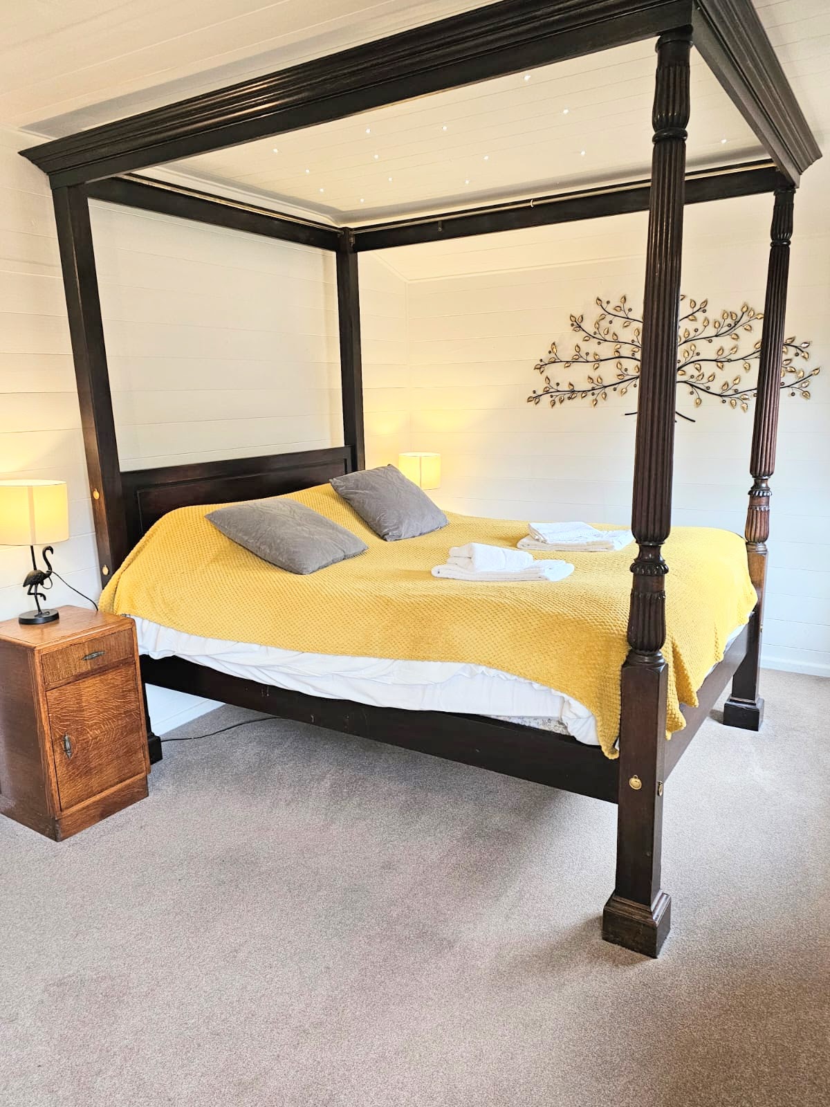 A four-poster bed is featured with a soft yellow throw and two grey pillows. Two bedside lamps provide gentle illumination beside a wooden nightstand. Warm light enhances the cozy ambiance within the room, complemented by a decorative wall accent above the bed.