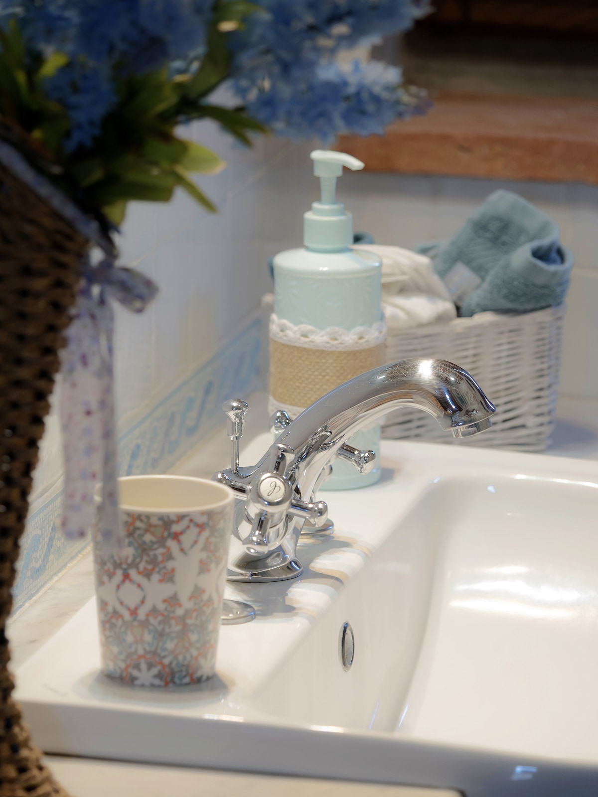 A bathroom sink area is displayed, featuring a polished silver faucet and decorative container holding liquid soap. A floral-patterned cup rests near the sink, alongside neatly arranged towels and a woven basket, contributing to a tidy atmosphere.
