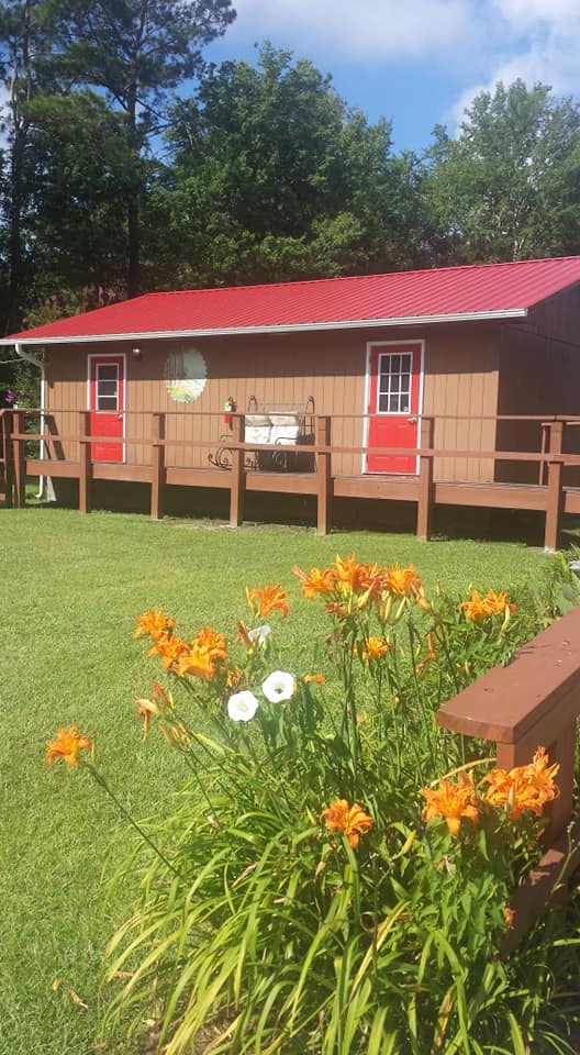 A charming cottage is surrounded by vibrant orange and white flowers in a well-maintained lawn. The building features a wooden deck with a red metal roof, complemented by two red doors and a circular decorative element on the side.