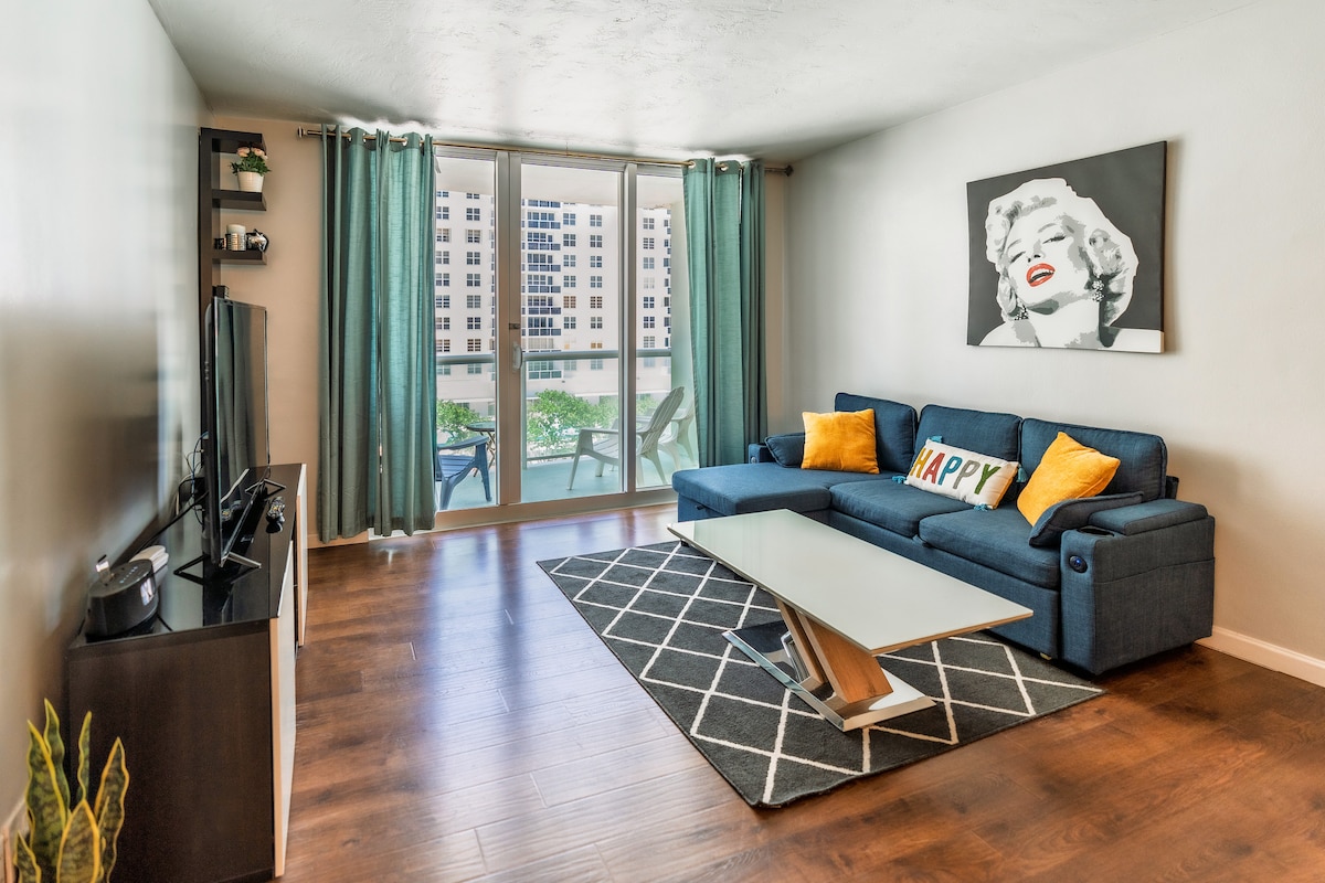 Moderná obývacia izba s balkónom a výhľadom na pláž, štýlová modrá pohovka, inteligentný televízor a veľké okná na prirodzené svetlo.