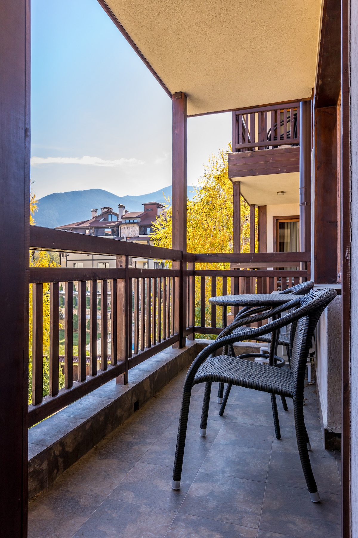 A private balcony features two black wicker chairs, offering an inviting space to enjoy the view. Surrounding trees showcase vibrant autumn colors, and distant mountains are visible under a clear blue sky. The balcony is framed by wooden railings, enhancing the outdoor experience.