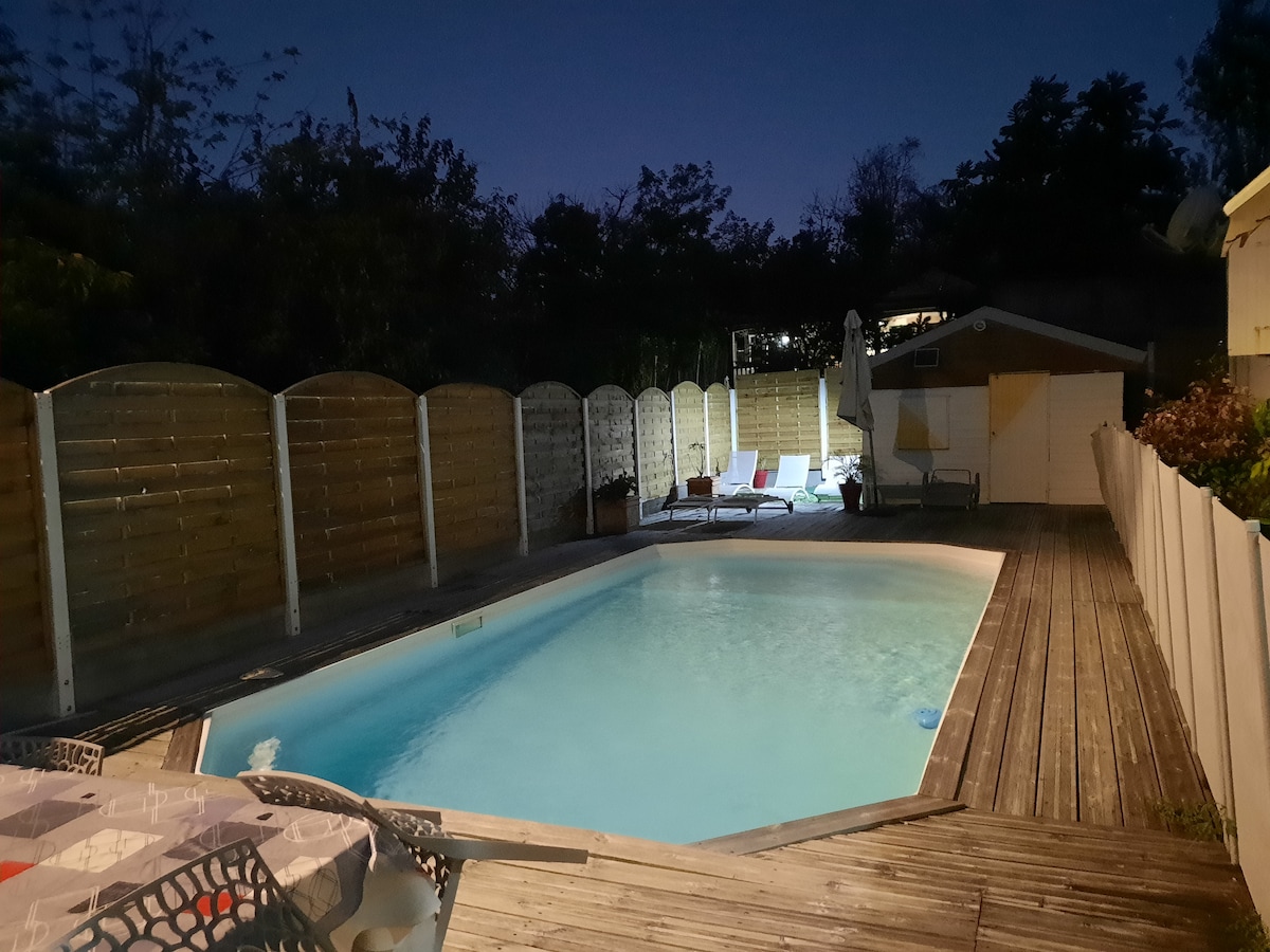 A tranquil swimming pool is illuminated at night, with calm water reflecting ambient lighting. Surrounding the pool, wooden decking creates a spacious area, while sun loungers are arranged for relaxation. A wooden fence provides privacy, with trees softly silhouetted against the evening sky.