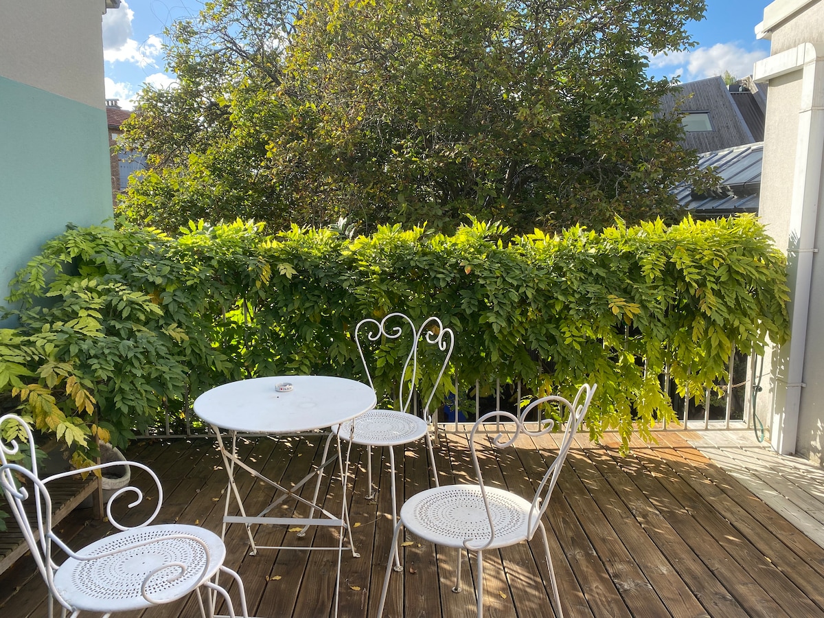 A serene outdoor terrace is presented, featuring a small round table accompanied by four white wrought iron chairs. Lush greenery surrounds the area, creating a peaceful environment. Warm sunlight filters through the leaves, enhancing the inviting setting for relaxation.