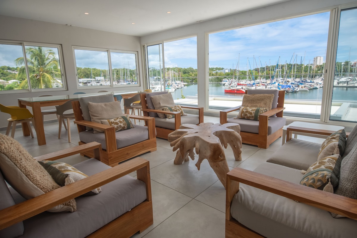 A spacious living area is surrounded by large windows, offering views of the marina and boats. Natural light fills the space, highlighting the wooden furniture with soft cushions. A unique wooden coffee table enhances the setting, creating an inviting atmosphere for relaxation.