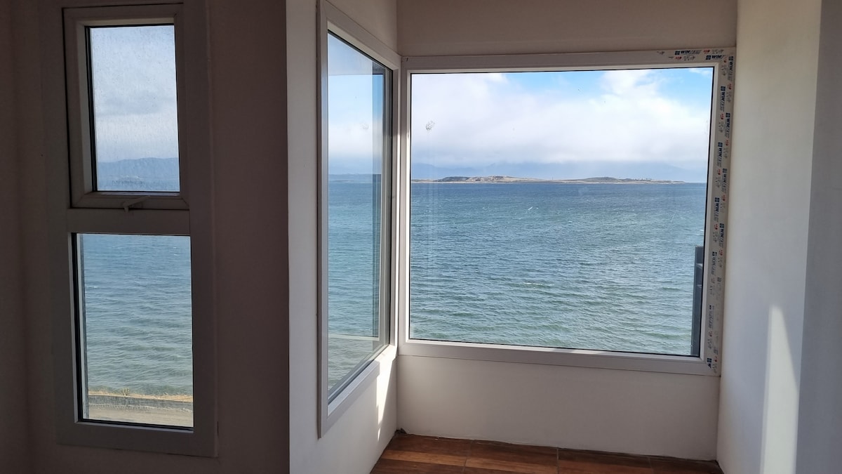 A bright corner of the room presents large windows offering expansive views of the calm water and distant shoreline. Natural light fills the space, highlighting the minimalist design. The peaceful blue hues of the water create a serene backdrop.