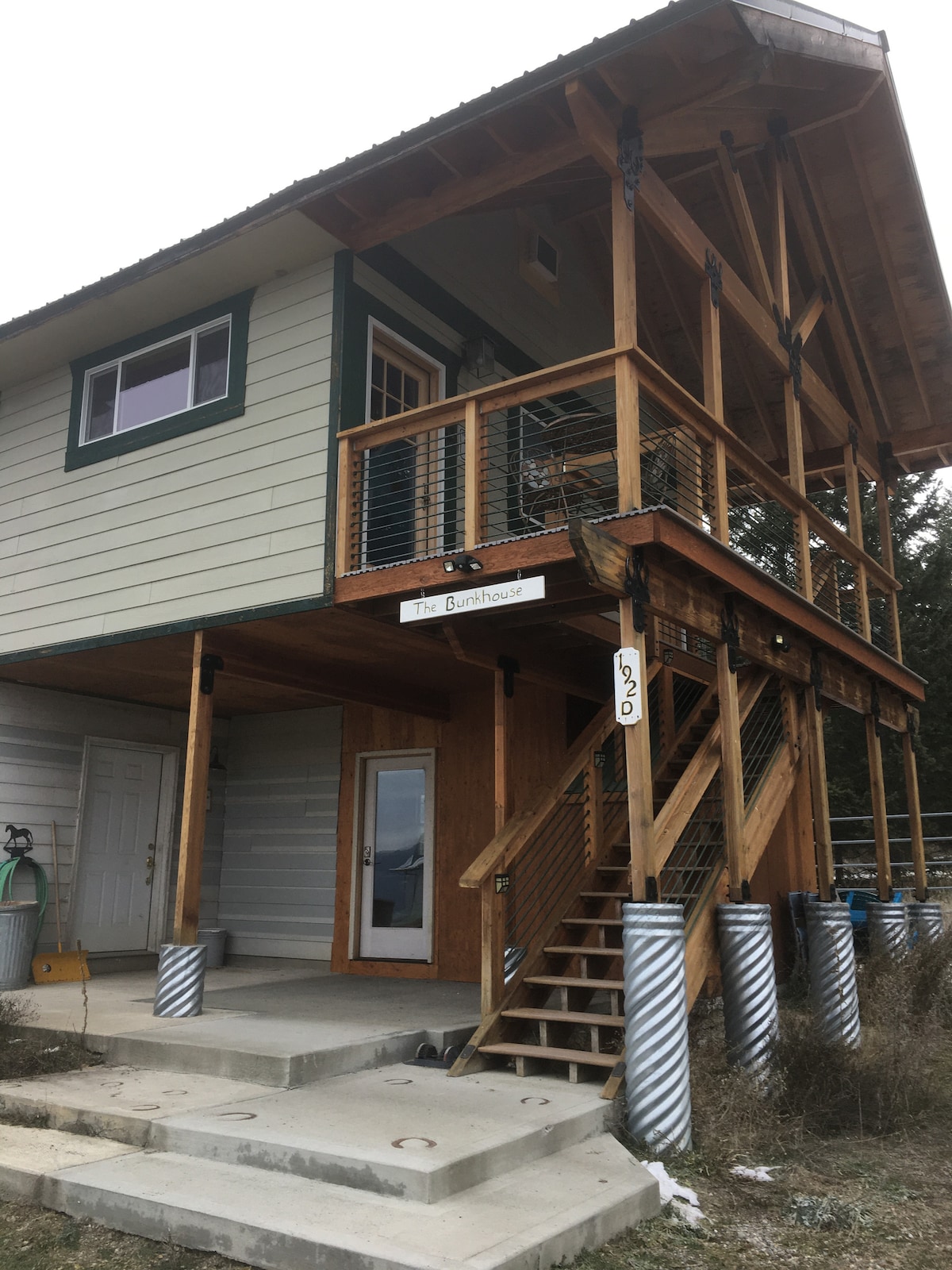 The exterior of the Bunkhouse is showcased under a cloudy sky. A wooden staircase leads to the entrance, framed by a covered porch with railings. The building features a combination of wood and siding with large windows, providing ample light to the interior.
