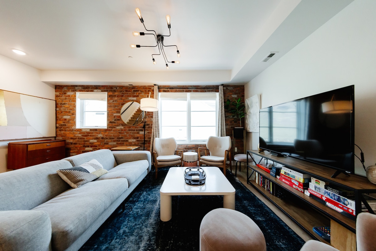 An open living space features a cozy sectional sofa against a textured brick wall. Natural light fills the room through two windows. A large TV is placed on a media shelf, showcasing board games and books. Accent chairs provide additional seating around a central coffee table.