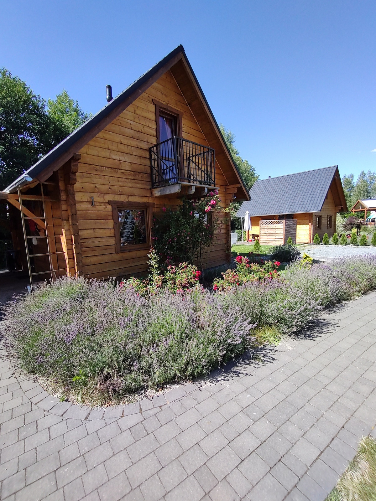 A charming wooden cottage is framed by colorful flowers and lush greenery. A small balcony is visible on the upper floor, overlooking a well-maintained path. The surrounding area features additional structures with gabled roofs against a clear blue sky.