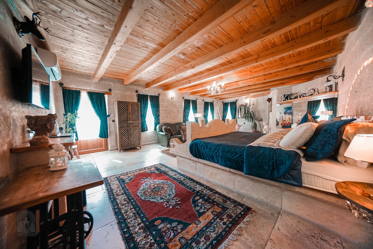 A spacious room features a large, comfortable bed with a decorative headboard and cozy bedding. Natural light filters through turquoise curtains, enhancing the warm interior. A stylish seating area is present, along with a wooden table and a traditional area rug complementing the stone walls.