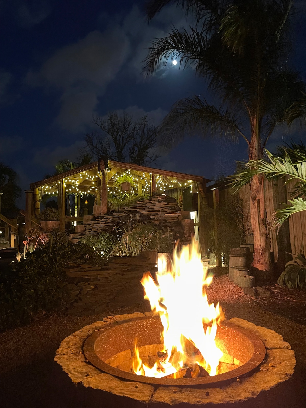 A warm glow from a fire pit illuminates the backyard, surrounded by lush greenery and palm trees. A deck adorned with string lights can be seen in the background, under a night sky featuring soft clouds and a bright moon.