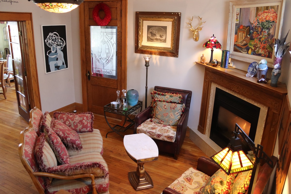 A cozy sitting area is showcased with patterned sofas and armchairs arranged around a marble-topped coffee table. Soft lighting is provided by a lamp and wall sconces, while a decorative fireplace adds warmth. Original art complements the historic wooden details.