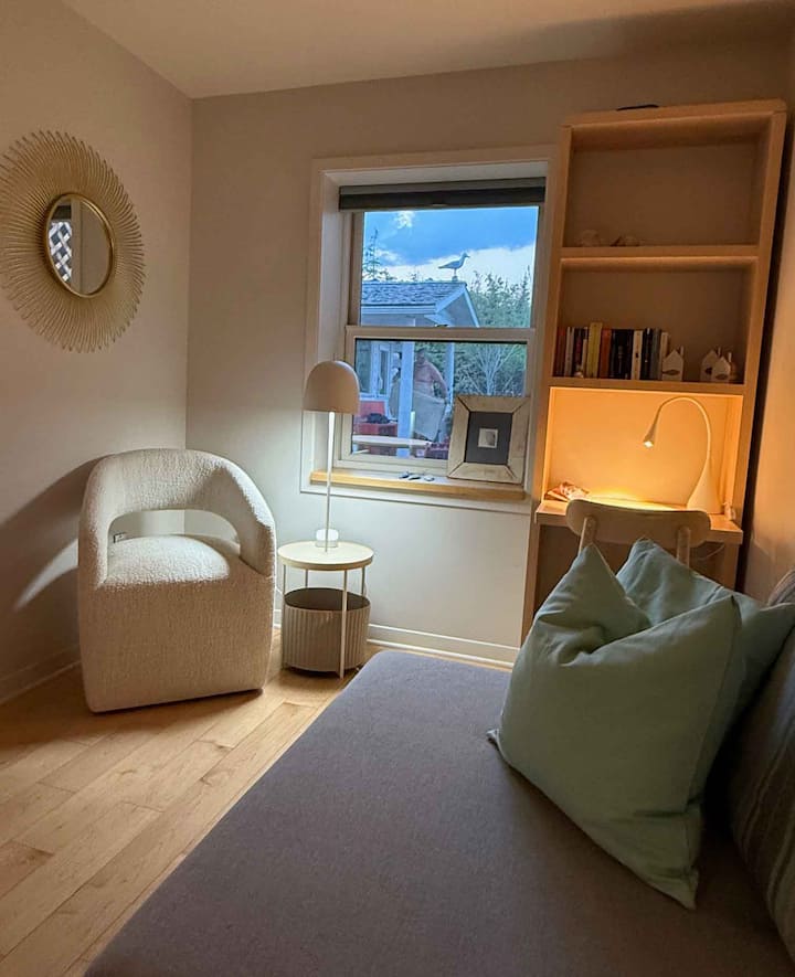 Cozy little room with a light-colored armchair, desk, and window with a view outside.