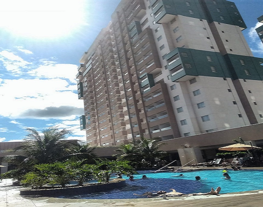 A spacious outdoor pool area is captured, surrounded by palm trees and lounge chairs. The multi-story building stands nearby, reflecting sunlight. Guests can be seen enjoying the water under a partly cloudy sky, showcasing a relaxed atmosphere.