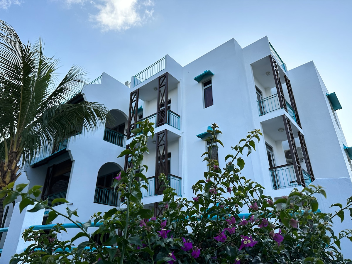 The image showcases a modern, multi-level building with white walls and turquoise accents. Lush greenery, including palm trees and flowering plants, frames the structure, which features multiple balconies with wooden railings and large windows that allow natural light to enter.