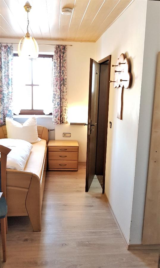 A well-lit single room is presented, featuring a cozy bed with decorative pillows and a wooden bedside table. Floral curtains frame a window, while a wooden door leads to the hallway. Soft wood flooring adds warmth to the space.