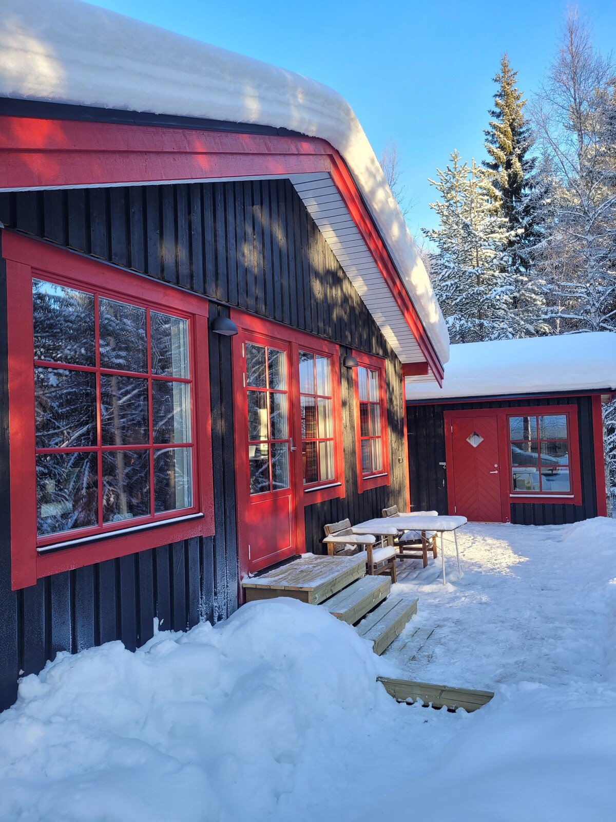 The exterior of a newly renovated cabin is shown, featuring a black façade with red accents, including windows and a door. Snow covers the ground and a small wooden deck leads up to the entrance, surrounded by trees blanketed in white.