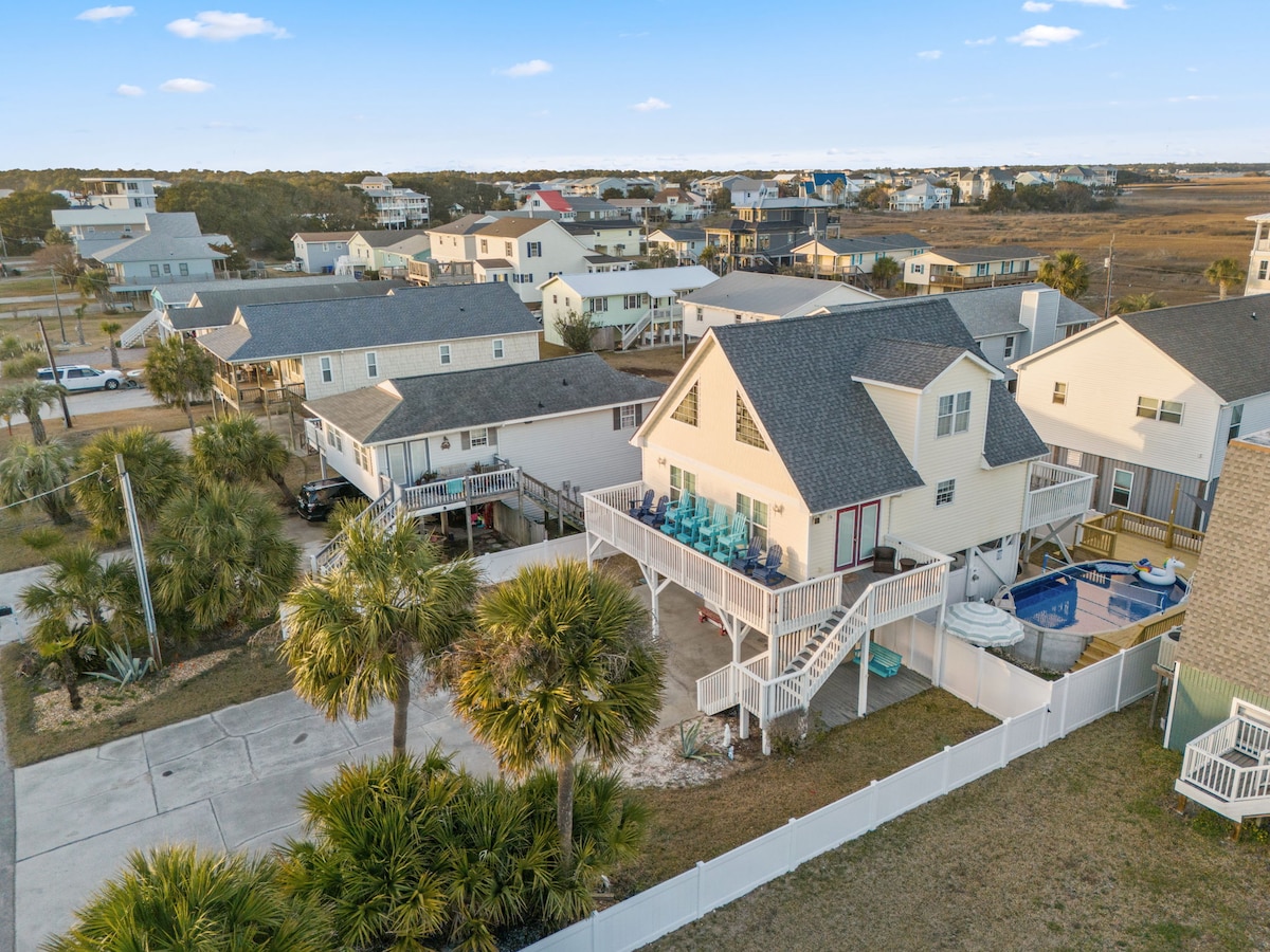An aerial view reveals a two-story house surrounded by a spacious yard with palm trees. A large deck extends from the back of the home, furnished with several blue chairs. Nearby streets and neighboring homes are visible in the background.