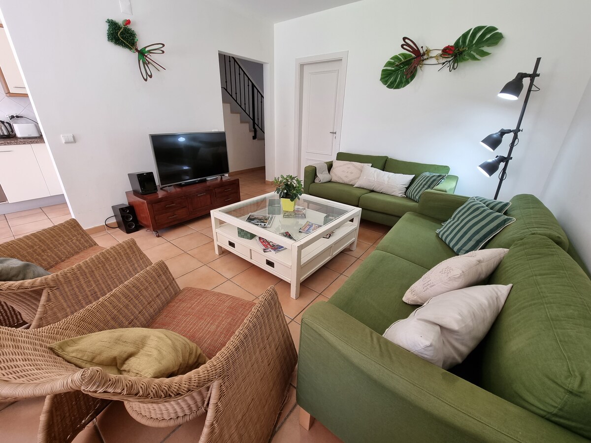 A welcoming living area features two green sofas and two wicker chairs arranged around a coffee table. A television sits against the wall, while decorative plants adorn the space. Natural light enhances the neutral tones of the room, providing a comfortable gathering spot.