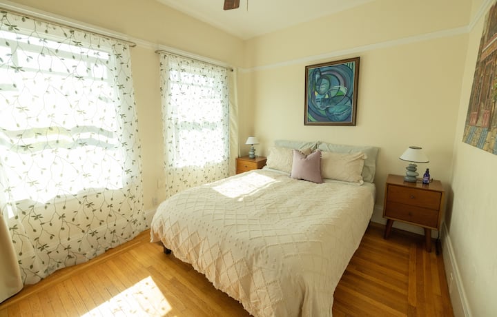 Enjoy a restful stay in this sunlit bedroom featuring warm wood floors, charming artwork, and soft, inviting bedding
