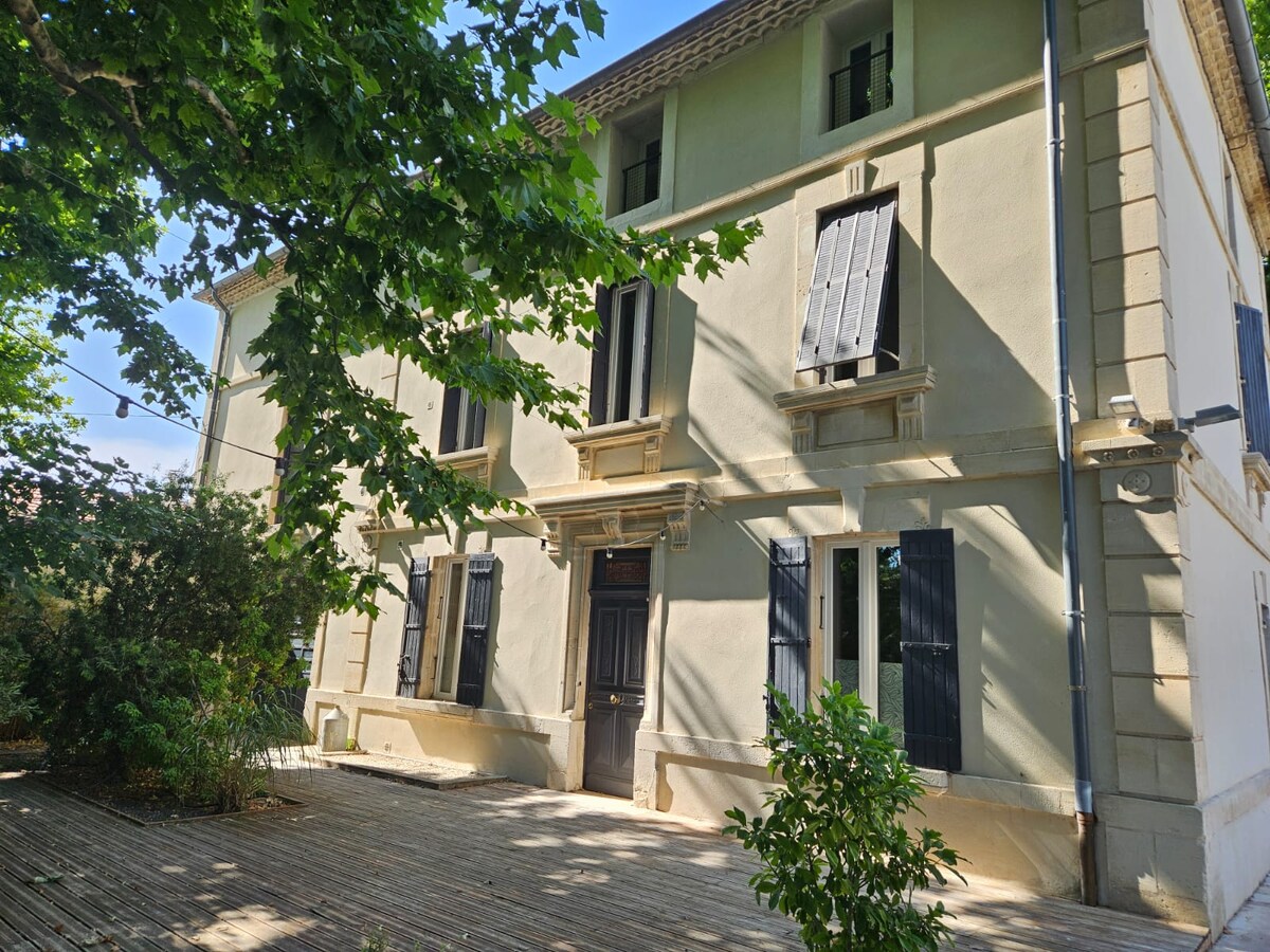 The exterior of a restored 19th-century building is shown, featuring light-colored walls and black shutters. A wooden deck area surrounds the entrance, while trees provide shade and greenery. The building's classic architectural elements, such as the entrance door and window arrangements, are prominently displayed.