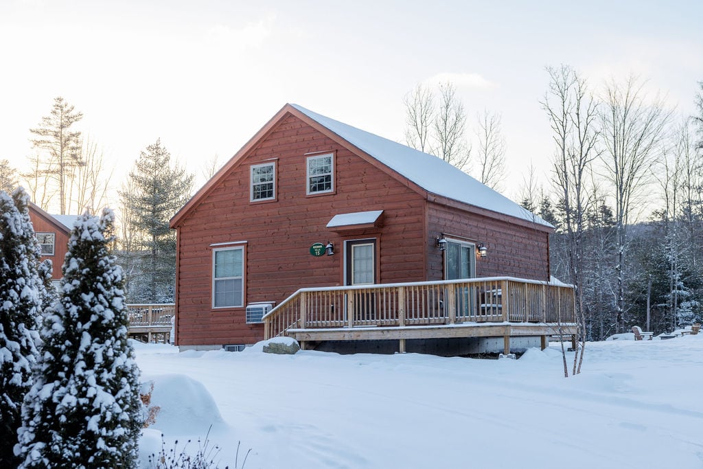 A cozy log cabin features an inviting wooden exterior, framed by a snow-covered landscape. The porch extends along the front, providing access to the indoors. Frosted windows reflect the soft light of dawn, while trees and snow enhance the serene winter setting.