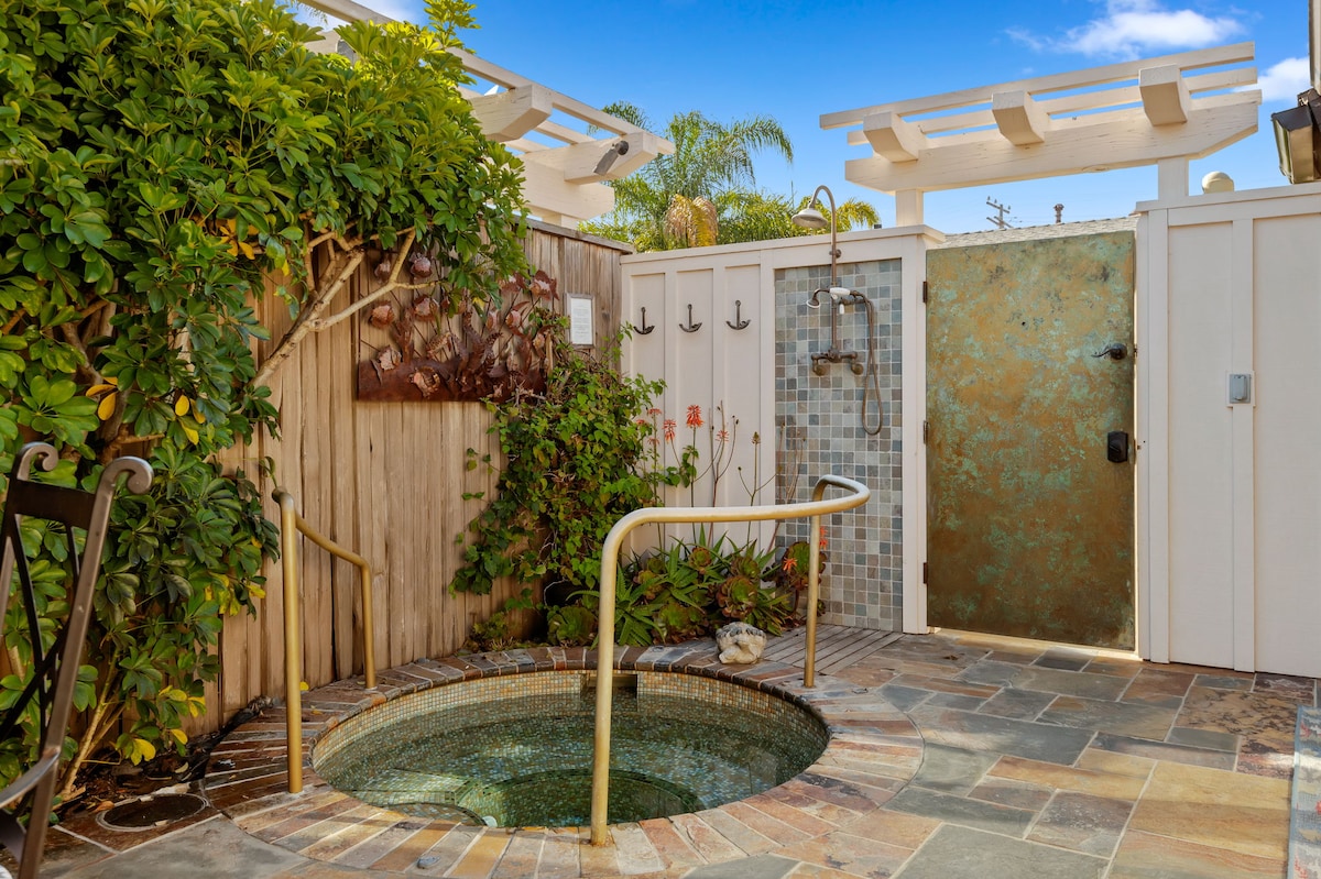 A private outdoor space features a built-in spa surrounded by natural stone tiles. Lush greenery adorns the area while a soothing outdoor shower stands nearby. The space is enclosed by wooden fencing and a partially transparent door, providing a serene atmosphere for relaxation.