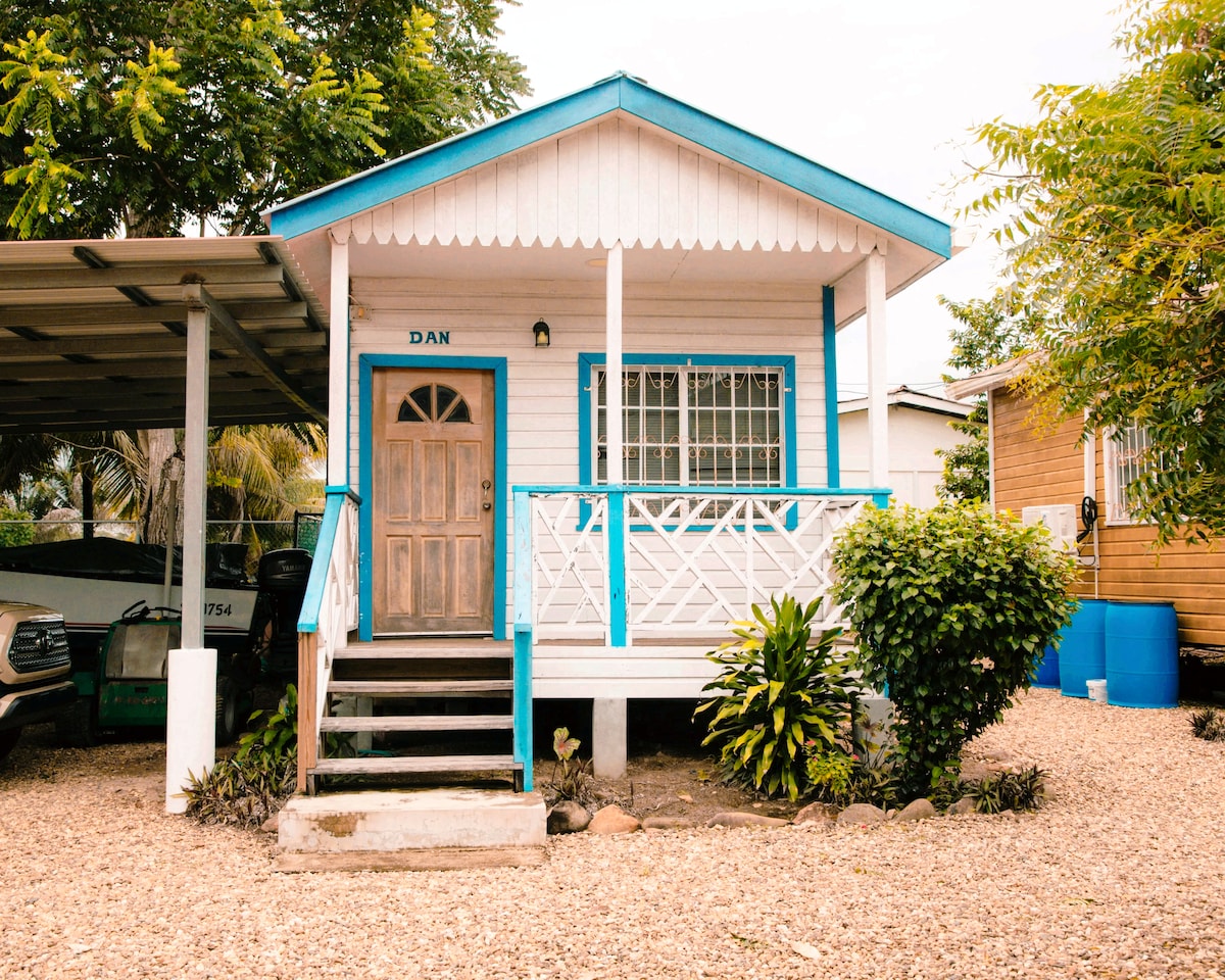 A charming cabana features a light blue exterior with a decorative roof overhang. Steps lead up to the entrance, which is highlighted by a wooden door. Lush greenery borders the front, while a gravel driveway accommodates vehicles.