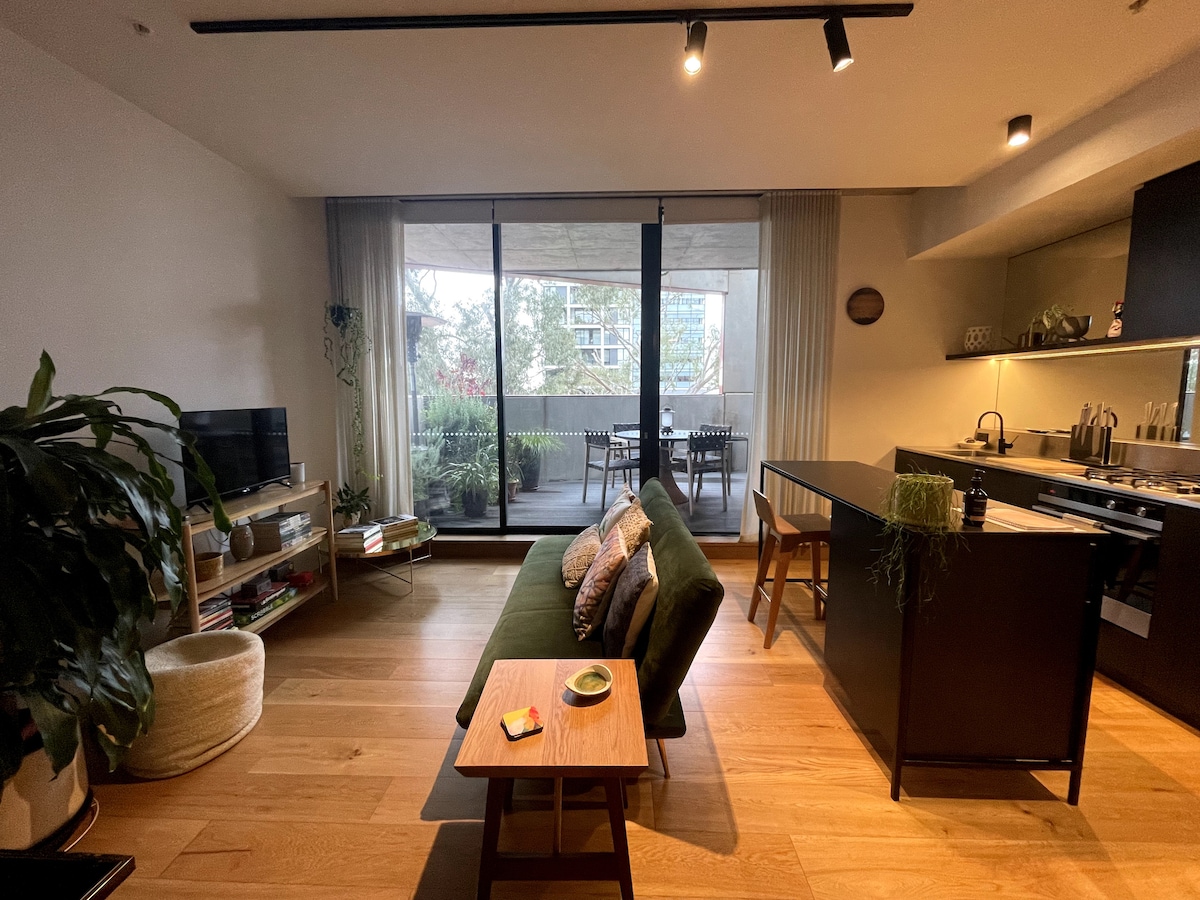 A modern living area is highlighted by a comfortable green sofa and a sleek wooden coffee table. Natural light fills the space from large glass doors, which overlook an outdoor balcony. An open kitchen area with minimalist design elements is also visible.
