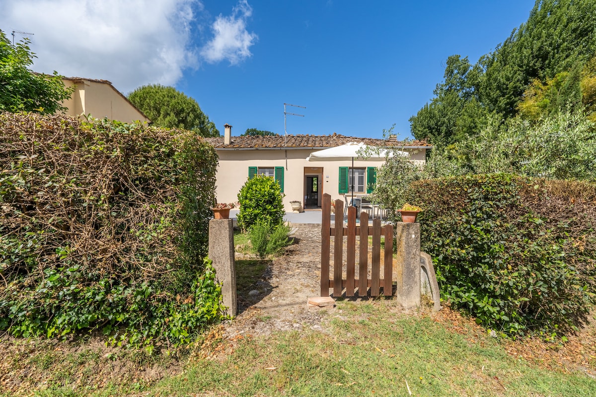 A welcoming entrance is framed by neatly trimmed hedges and a wooden gate, leading towards a single-story building with traditional architecture. Greenery surrounds the property, offering a sense of privacy and tranquility. Blue skies and clouds contribute to a serene outdoor atmosphere.